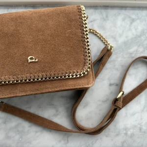 Brun mocka axelväska med kedjedetaljer - Snygg brun axelväska i mocka med guldfärgad kedja längs kanterna och axelremmen. Väskan har en magnetknapp för stängning och flera fack inuti, inklusive ett med dragkedja. Helt ny!