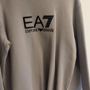 Grå tröja från Emporio Armani - Snygg grå tröja från Emporio Armani med EA7-logga på bröstet. Perfekt för en stilren och avslappnad look. Tröjan har långa ärmar och en klassisk passform.