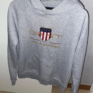 Grå hoodie från GANT - Säljer en stilren grå hoodie från GANT med broderad logga och text 'American Sportswear' på framsidan. Tröjan har en klassisk känguruficka och en bekväm huva. Perfekt för en avslappnad stil.