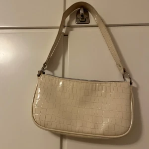 Beige axelväska med krokodilmönster - Snygg beige axelväska med krokodilmönster och dragkedja. Perfekt för att ge en stilren touch till din outfit. Väskan har en enkel design med ett kort axelband.