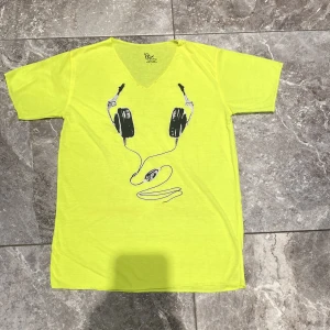 Gul t-shirt med hörlursmotiv från Peachy - Säljer en cool gul t-shirt från Peachy med ett tryck av hörlurar på framsidan. T-shirten har en v-ringad hals och är kortärmad. Perfekt för en avslappnad stil. Köpt i Thailand 