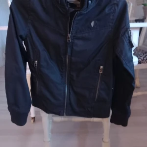 Svart bomberjacka med dragkedja - Snygg svart bomberjacka med dragkedja framtill och praktiska fickor. Jackan har en klassisk design med ribbade muddar vid ärmslut och nederkant. Perfekt för en cool och avslappnad stil.