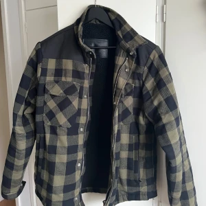Mil-Tec jacka - Säljer en snygg rutig jacka med fleecefoder. Jackan har en dragkedja framtill och två bröstfickor med knappar. Den är perfekt för kyligare dagar och har en klassisk look med sina gröna och svarta rutor. Även mycket slitstark. 