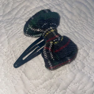 Rutig rosett hårklämma - Söt hårklämma med en rutig rosett i grönt, rött, blått och vitt. Perfekt för att ge en färgglad touch till din frisyr. Rosetten är fäst på en plastklämma som håller håret på plats.