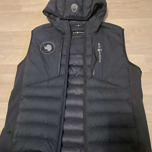 Sail Racing väst - Snygg svart dunväst från Sail Racing med dragkedja och huva. Västen har en stilren design med logotypdetaljer på bröstet och huvan. Perfekt för kyligare dagar. Använd i ungefär ett år. Finns inga fel. Nypris ca. 2000