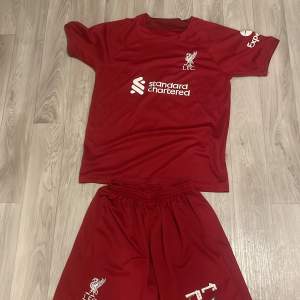 Säljer en röd Liverpool fotbollströja med matchande shorts. Tröjan är kortärmad med klubbens logga och sponsortryck på framsidan. Perfekt för alla Liverpool-fans som vill visa sitt stöd. Setet är i bra skick och redo för nästa match eller träning! Darwin Nunez 