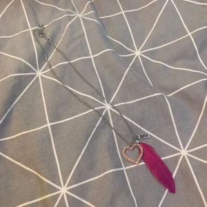 Halsband med hjärta och fjäder - Säljer ett superfint halsband med ett glittrigt hjärthänge och en rosa fjäder. Perfekt för att ge till din BFF! Kedjan är i silverfärg och har en liten bricka med texten 'BFF'. Man kan ta bort fjärden och bff om man bara vill ha hjärtat. 