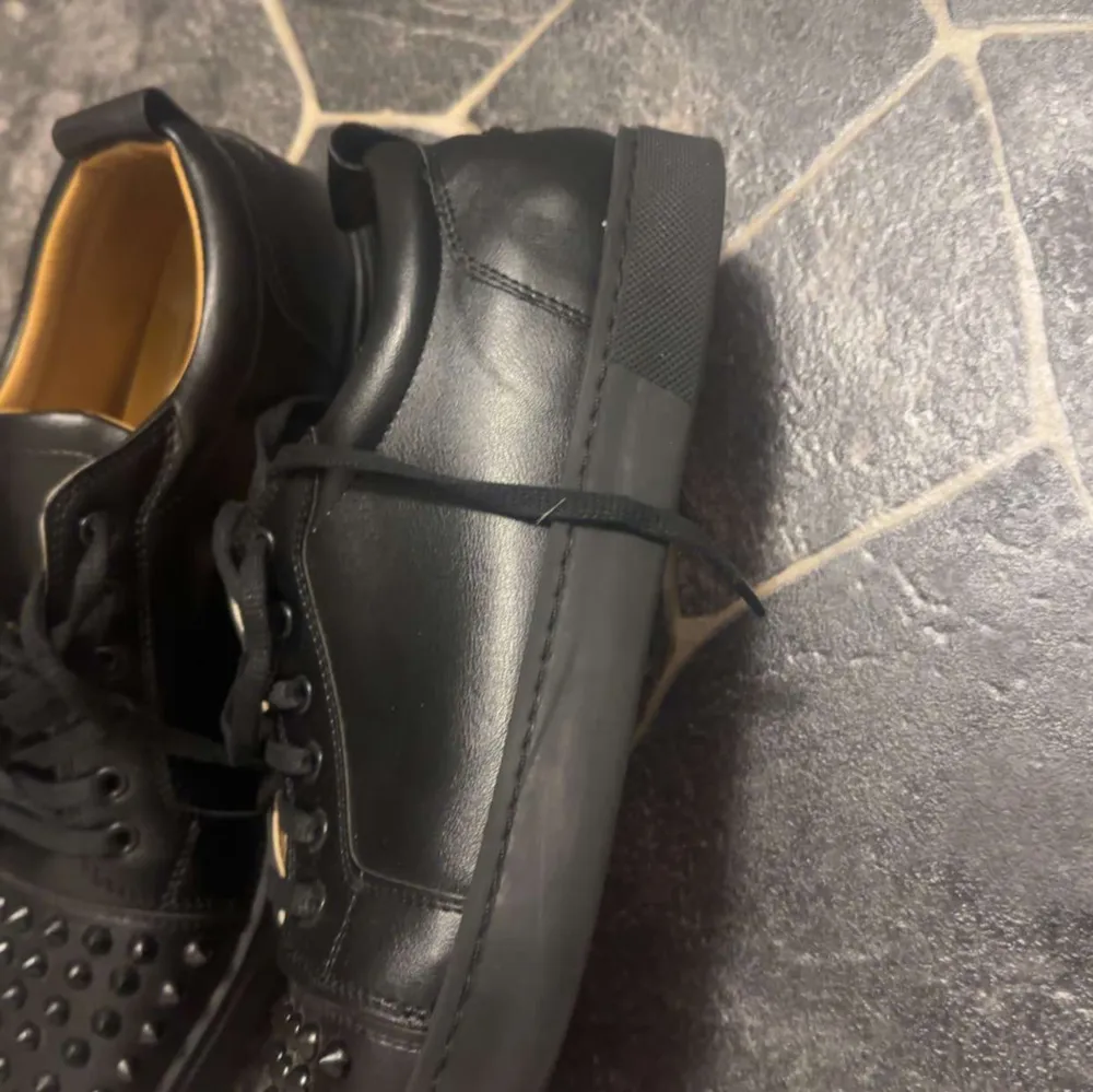 Säljer ett par svarta sneakers från Christian Louboutin i skinn med coola nitar på tån. De har den klassiska röda sulan och snörning framtill. Perfekta för att ge din outfit en edgy touch! Använda men i bra skick.. Skor.