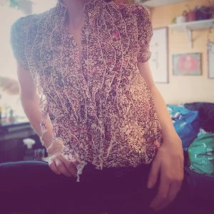 Blommig blus med volanger - Säljer en superfin blommig blus med volanger. Den har ett färgglatt mönster i rosa, svart och beige. Perfekt för både vår och sommar! Blusen har knappar framtill och är gjord i ett lätt och luftigt material. Passar perfekt till både jeans och kjol för en söt och chic look.