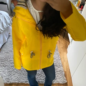 Gul regnjacka med randig huva - Säljer en superfin gul regnjacka med randigt foder i huvan. Den har dragkedja och snörning i midjan för bästa passform. Perfekt för regniga dagar och vårpromenader. Jackan är i bra skick och redo för nya äventyr! 🌧️💛