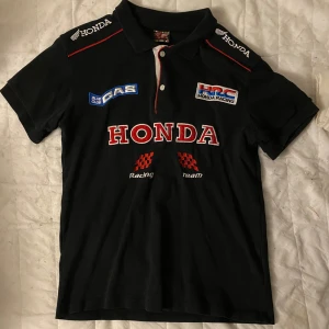Svart Honda Racing pikétröja - Säljer en svart Honda Racing pikétröja med korta ärmar. Den har flera broderade logotyper, inklusive Honda och HRC, både fram och bak. Perfekt för racingfans! Tröjan är i bra skick och passar till både vardag och event.
