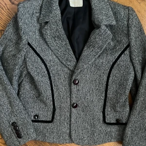 Grå kavaj med svarta detaljer - Säljer en snygg vintage  grå kavaj perfekt för hösten  . Den har en klassisk krage och knäpps med två knappar framtill. Ärmarna är långa och har dekorativa knappar vid ärmsluten. Storlek är 44 ,översize jättefin men polotröja under 