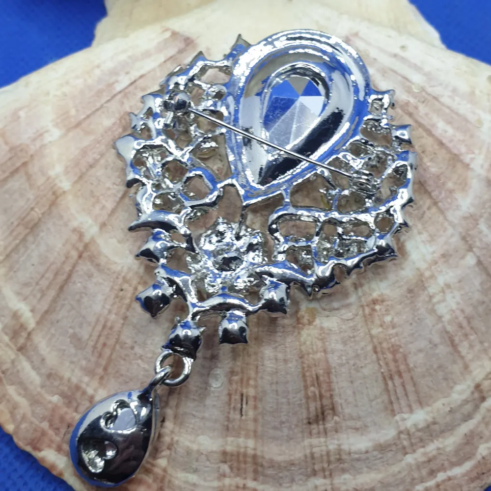 En brosch föreställande en Rojalistisk Kristallbrosch. Storlek ca 10x6cm. Silverfärgad metall med vacker ytstruktur och ytan täckt av kristaller I olika storlekar och former, en kristall droppe hänger undertill. Grymt snygg brosch.. Asusteet.