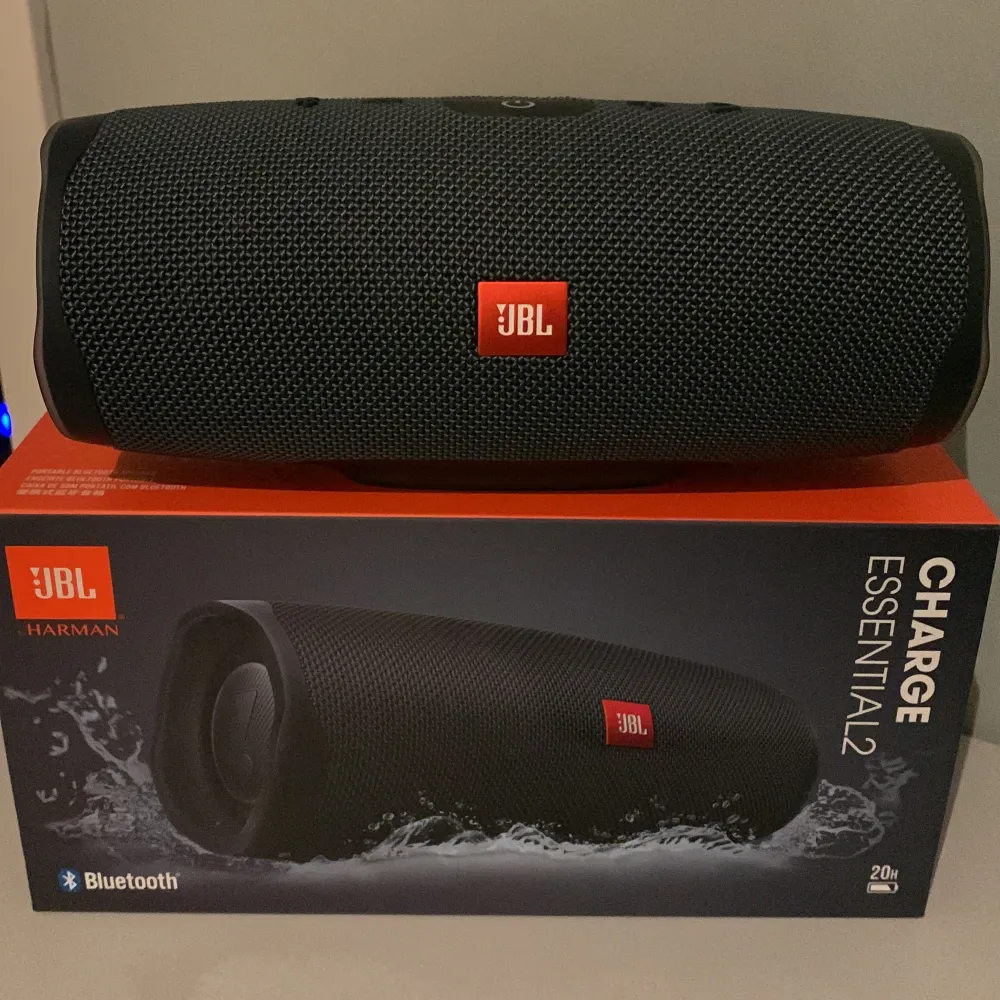 Säljer en JBL Charge Essential 2 Bluetooth-högtalare i nyskick. Den har en snygg och robust design med ett mörkt tygöverdrag och den klassiska JBL-loggan i rött. Perfekt för att ta med på äventyr eller bara ha hemma för att njuta av grymt ljud. Högtalaren är vattentålig och har lång batteritid, vilket gör den idealisk för både inomhus- och utomhusbruk. Kommer med originalförpackning.. Accessoarer.