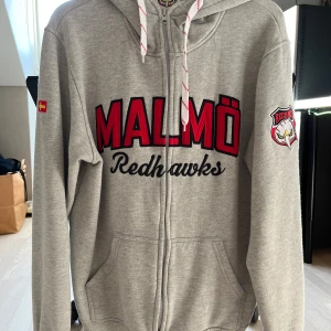 Grå hoodie från Malmö Redhawks - Säljer en grå hoodie från Malmö Redhawks i storlek L. En hoodie för den som verkligen vill stödja sitt lag. Har den röda örnen och skåneflaggen på båda armarna.