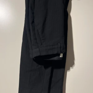 Svarta kostymbyxor med diskreta ränder - Säljer ett par stiliga svarta kostymbyxor med diskreta ränder. Perfekta för både formella tillfällen och kontoret. Byxorna har en klassisk passform och är gjorda i ett bekvämt material. De har en snygg detalj med uppvikta benslut. Storlek 40