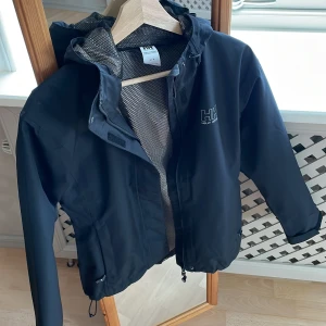 REGNJACKA  - Svart regn/vindjacka från HellyHansen, endast använd ett fåtal gånger pga för liten så den är som ny! Dragsko nertill. Upplever den liten i storlek.