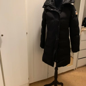 Svart dunjacka från Carhartt - Säljer en svart lång dunjacka från Svea . Den är perfekt för kalla vinterdagar med sin långa design och varma fyllning. Jackan har en huva och stängs med både dragkedja och knappar. Den är i nyskick och har långa ärmar. Perfekt för vintern!