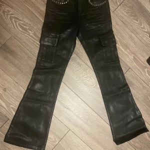 Svarta Vendetta waxed jeans från Vengeance Studios - Säljer ett par svarta  Vendetta waxed jeans  från Vengeance Studios. Byxorna har en cool broderad design på bakfickorna och är dekorerade med nitar. De har flera praktiska fickor. Perfekta för en edgy look!