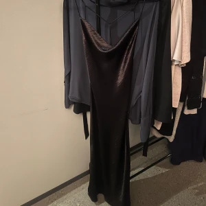 Svart glansigklänning med öppen rygg - Elegant svart långklänning med en stilren design. Klänningen har en öppen rygg med tunna band som korsar varandra, vilket ger en sofistikerad look. Perfekt för speciella tillfällen.