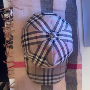 Rutig keps från Burberry - Snygg rutig keps från Burberry i klassiskt mönster med beige, svart och röd färg. Kepsen har en justerbar rem baktill och en broderad Burberry-logga. Perfekt för en stilren look. Skick 9-10 pris kan diskuteras 