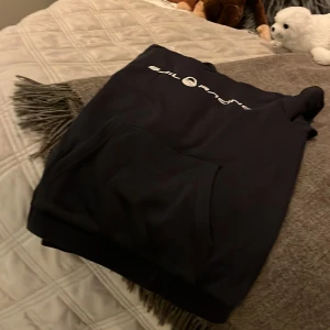 Svart hoodie från Sail Racing - Säljer en svart hoodie från Sail Racing med vit logga på bröstet. Den har en klassisk känguruficka framtill och är perfekt för en avslappnad stil. Passar bra till både vardag och chill.