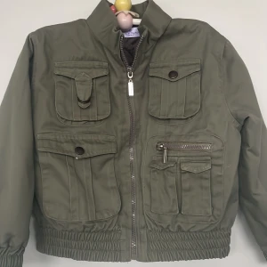 Olivgrön jacka från Naf Naf Enfant st.110 - Snygg olivgrön jacka med flera praktiska fickor och dragkedja framtill st.110. Jackan har en quiltad insida för extra komfort och stil. Perfekt för höst och vår.