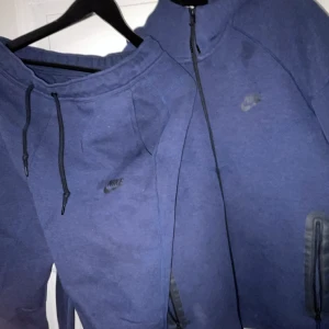 Blå hoodie och byxa  från Nike - Säljer en blå hoodie från Nike med dragkedja och justerbar huva. Och ett par byxor Perfekt för en avslappnad stil. Den har en diskret Nike-logga på bröstet och låret och dem är tillverkad i ett mjukt material.
