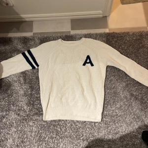 Vit stickad tröja med bokstaven A - Säljer en vit stickad tröja med en mörkblå bokstav A på bröstet och två mörkblå ränder på ena ärmen, perfekt för kyligare dagar.