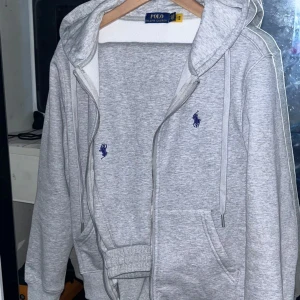 Grå dress från Ralph Lauren - Säljer en stilren grå hoodie och byxor  från Ralph Lauren med dragkedja och huva. Tröjan har det klassiska Ralph Lauren-logotypen broderad på bröstet. Perfekt för en avslappnad look.