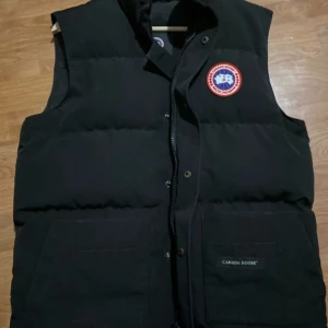 Svart dunväst från Canada Goose - Säljer en svart dunväst från Canada Goose med deras ikoniska märke på bröstet. Västen har en dragkedja och knappar framtill samt två stora fickor. Perfekt för kyligare dagar.
