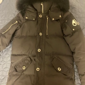 Svart dunjacka från Moose Knuckles - Säljer en svart dunjacka från Moose Knuckles med pälskantad huva och flera praktiska fickor. Jackan har dragkedja och knappar framtill samt en cool patch på insidan. Perfekt för kalla dagar! 🖤