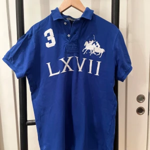 Blå pikétröja från Ralph Lauren - Snygg blå pikétröja från Ralph Lauren med broderad logga och siffran 3 på bröstet. Tröjan har en klassisk krage och knappar framtill. 