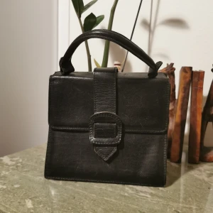 Svart kvadratisk handväska med spänne - Svart kvadratisk läder handväska med spänne. Det är en dragspelsväska (se sista bilden) så rymligare än man tror. Flera fack och lagom med slitage🖤. 