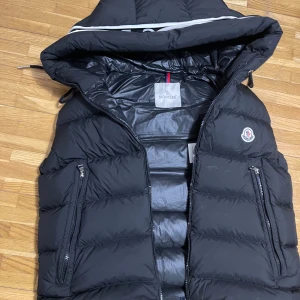 Svart väst från Moncler - Snygg svart väst från Moncler med huva och dragkedja. Jackan har Moncler-logga på bröstet och en cool serietidningsdetalj på insidan. Perfekt för kyliga dagar. Den är i storlek 2/M