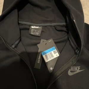 Svart hoodie från Nike - Säljer en stilren svart hoodie från Nike med dragkedja och huva. Perfekt för en casual look. Den har långa ärmar och en klassisk passform med fickor framtill.