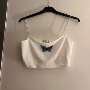 Vit linnetopp med fjärilsdetalj - Säljer en söt vit linnetopp från NLY One med tunna axelband och en blå fjäril på bröstet. Perfekt för varma sommardagar eller som ett lager under en jacka. Toppen är enkel men har en fin detalj som gör den unik.