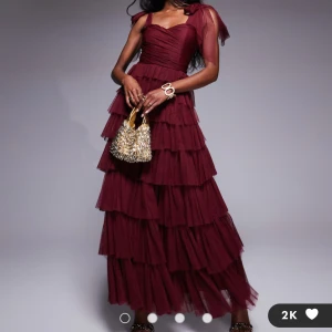 Vinröd långklänning med volanger - Elegant vinröd långklänning med flera lager av volanger och breda axelband. Perfekt för en glamorös kväll. Klänningen har en smickrande passform och en vacker struktur som ger en lyxig känsla.