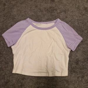 Vit och lila croppad t-shirt - Säljer en söt croppad t-shirt i vitt med lila raglanärmar. Perfekt för en avslappnad stil. T-shirten har en rund halsringning och korta ärmar.