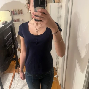 Mörkblå topp med knappar - Säljer en mörkblå topp med korta ärmar och knappar framtill. Perfekt för en avslappnad stil. Toppen har en enkel och stilren design.