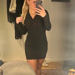 Svart klänning med knappar - Snygg svart klänning med knappar framtill och långärmad design. Den har en tight passform som framhäver figuren och en kort längd som ger en modern look. Perfekt för en kväll ute eller en speciell tillställning.