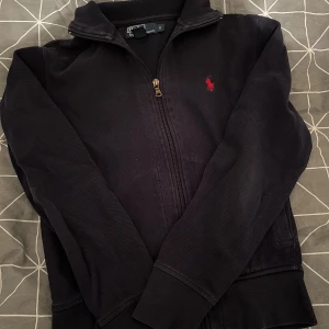 Mörkblå full zip från Polo Ralph Lauren - Säljer en stilren tröja från Polo Ralph Lauren med dragkedja och röd broderad logga på bröstet. Tröjan har långa ärmar och ribbade muddar. Perfekt för en casual look.