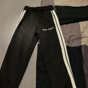 Svarta byxor från Palm Angels - Snygga svarta byxor från Palm Angels med vita sidoremsor och elastisk midja. Perfekta för en avslappnad stil med en touch av lyx. Märkesloggan är tryckt på framsidan. Självklart äkta, pris går att diskutera 