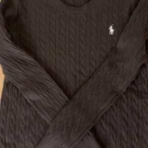 Svart kabelstickad tröja från Polo Ralph Lauren - Säljer en stilren svart kabelstickad tröja från Polo Ralph Lauren. Tröjan har en klassisk design med rund hals och det ikoniska broderade logotypen på bröstet. Perfekt för en elegant och avslappnad look.