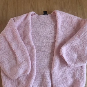 Rosa stickad kofta - Mysig rosa stickad kofta med en öppen design. Perfekt för att hålla sig varm under kyliga dagar. Koftan har en enkel och stilren look som passar till många olika outfits.