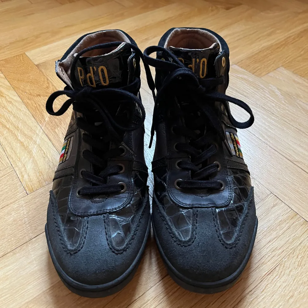 Snygga svarta sneakers från Pantofola d'Oro med krokodilmönstrade detaljer och färgglada emblem på sidan. Skorna har snörning och en högre ankel för extra stöd. Perfekta för en stilren look med en touch av unik design.. Kengät.