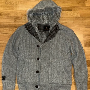 Grå stickad jacka med pälsfodrad huva och fodring - Säljer en grå stickad jacka  med en mysig pälsfodrad huva. Jackan har knappar framtill och ett rutigt mönster på insidan av huvan. Perfekt för kyliga dagar! Storlek M passar s med som ny