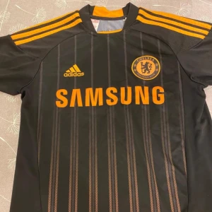 Svart Chelsea fotbollströja från Adidas - Snygg svart Chelsea fotbollströja från Adidas med orangea detaljer och ränder. Tröjan har korta ärmar och klubbens emblem på bröstet. Perfekt för fans som vill visa sitt stöd med stil! 🖤🧡