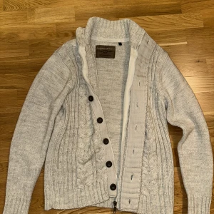 Grå stickad kofta cardigan beige - Säljer en stilren grå stickad kofta från Angelo Litrico. Koftan har ett flätmönster framtill och stängs med knappar. Den har en hög krage och långa ärmar, perfekt för kyligare dagar. Passar bra till både vardag och mer uppklädda tillfällen.