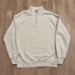 Polotröja med dragkedja - Säljer en stilren crèmefärgad tröja med halv dragkedja och hög krage. Tröjan har långa ärmar och ribbade muddar vid ärmslut och nederkant. Perfekt för en avslappnad look.